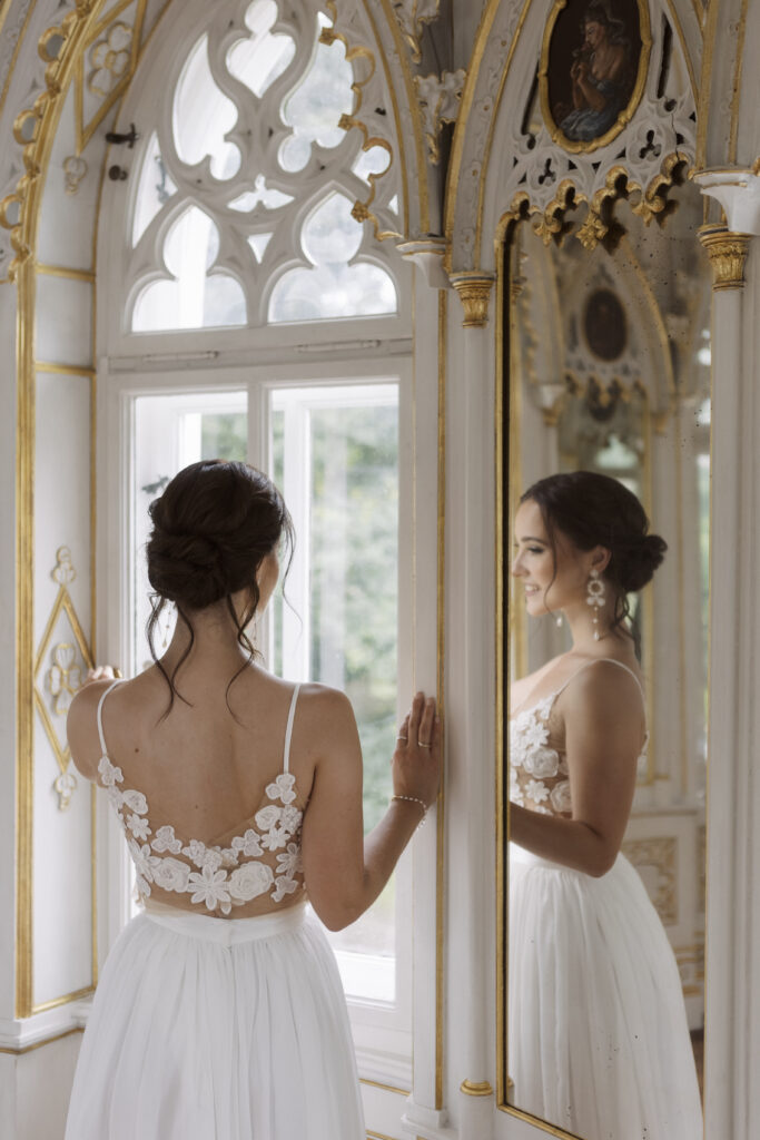 Eine Braut in einem weißen Kleid steht am Fenster und bewundert ihren Brautschmuck, während sie ihr Spiegelbild in einem verzierten Spiegel betrachtet.