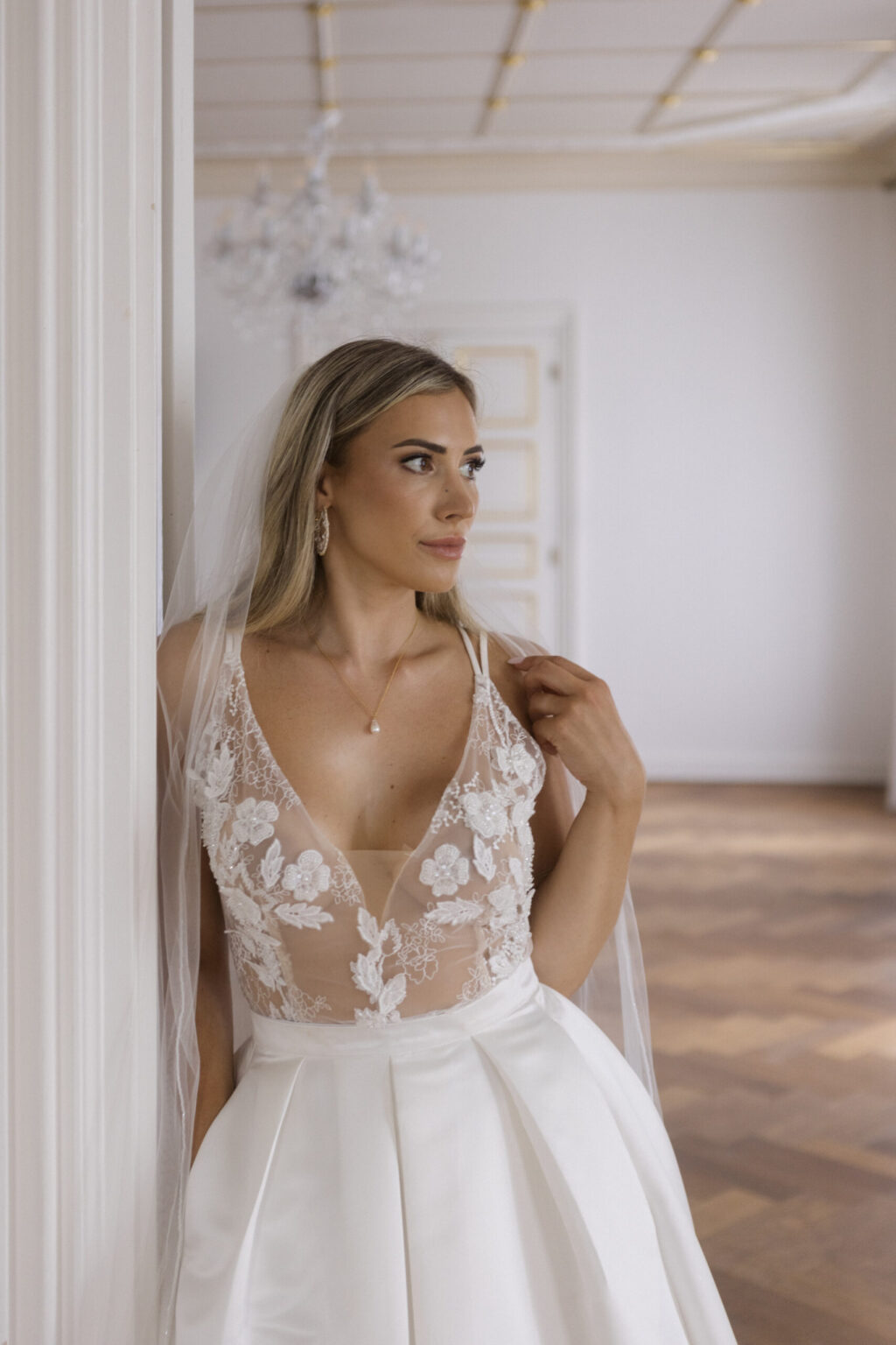 Eine Braut in einem weißen, mit Blumen verzierten Spitzenkleid von Elisa Florence steht in einem Raum und blickt nachdenklich zur Seite.