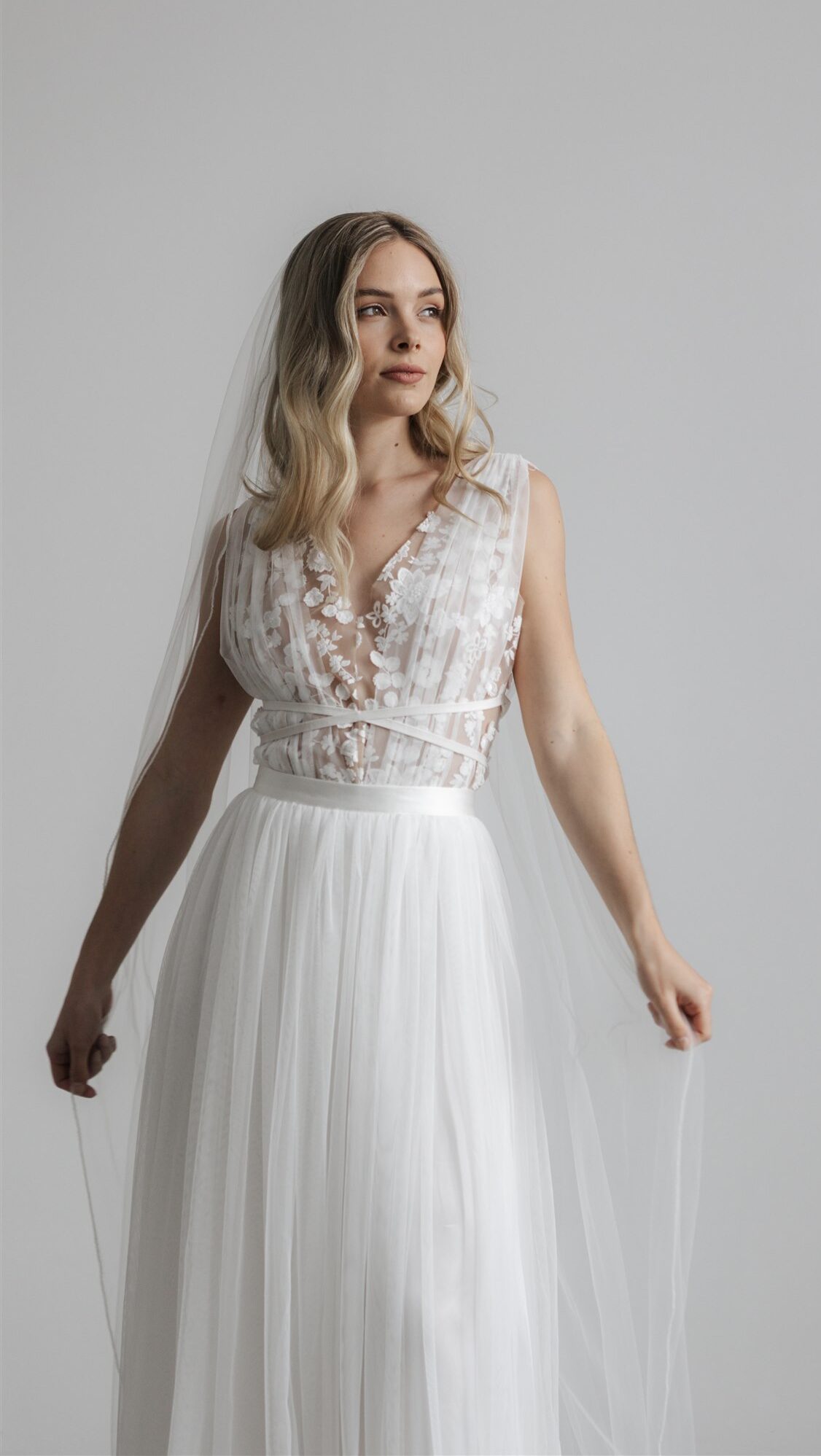 Eine Frau in einem weißen, ärmellosen Brautkleid mit floralen Details und einem Schleier, die vor einem grauen Hintergrund steht – eine elegante Hommage an die Brautmode, inspiriert von der zeitlosen Anmut von Felina Paris.