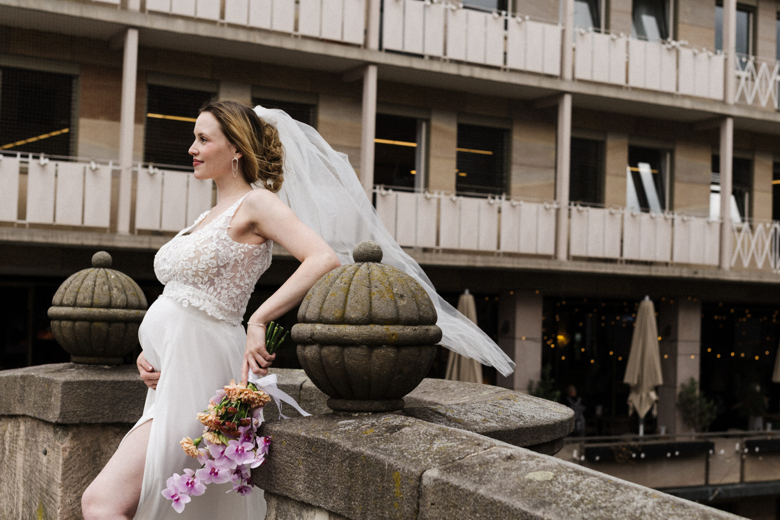 Eine schwangere Braut in weißem Kleid und mit Schleier hält einen Blumenstrauß in der Hand und steht an einem Steinbalustrade, hinter ihr ist ein Gebäude zu sehen.
