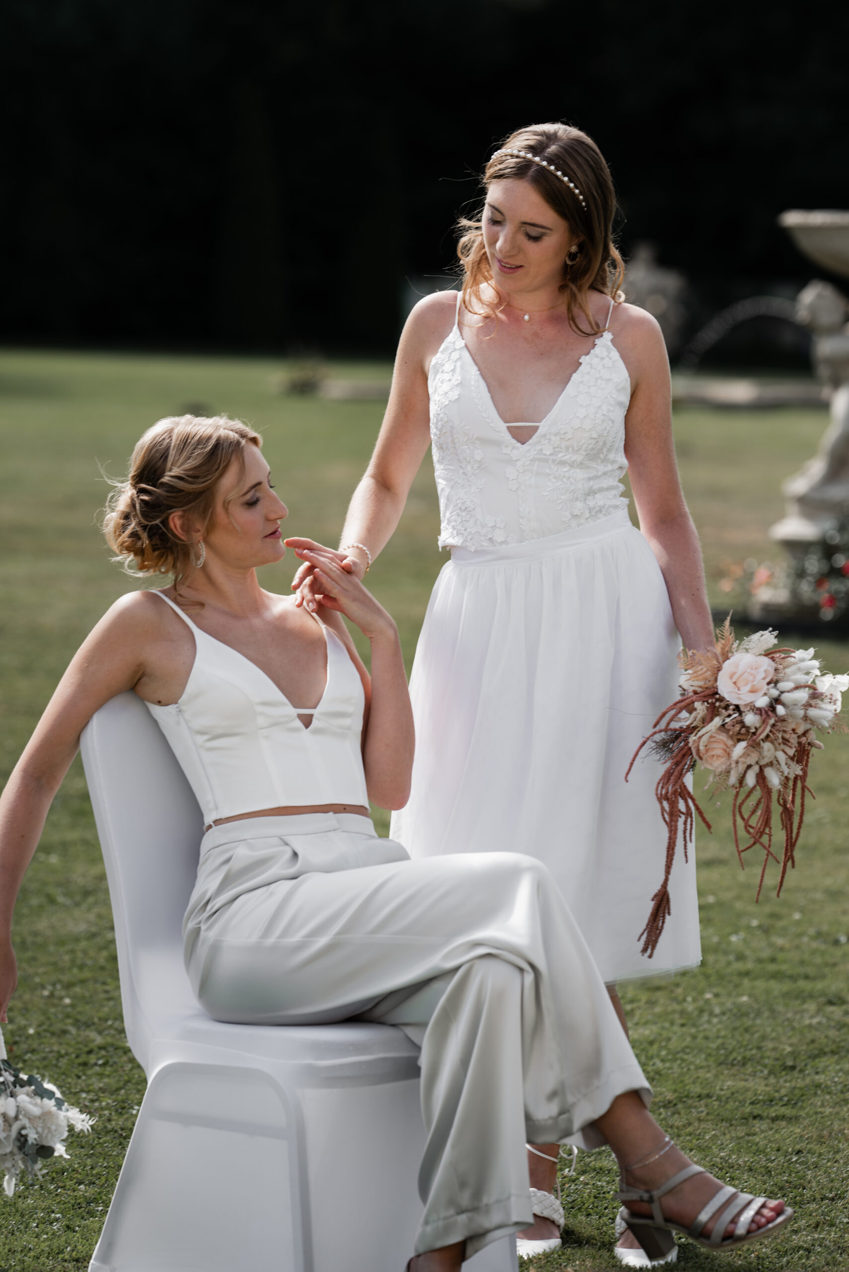 Zwei Frauen in weißen Brautkleidern, eine sitzt, die andere steht, genießen gemeinsam einen zärtlichen Moment im Freien, umgeben von Blumen.
