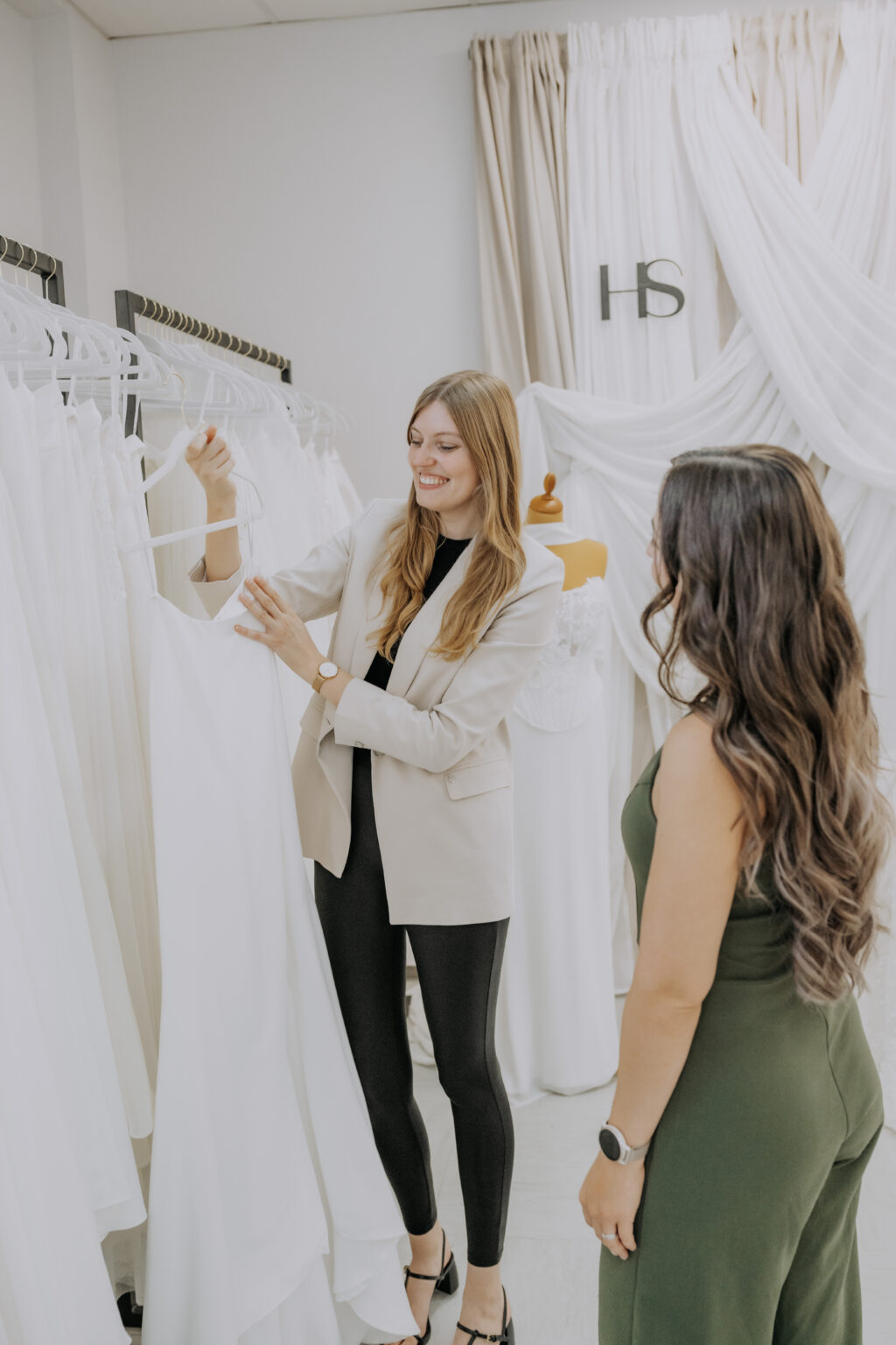 Zwei Frauen betrachten ein weißes Brautkleid in einem Brautmodengeschäft, in dem weiße Kleider an Kleiderständern hängen.