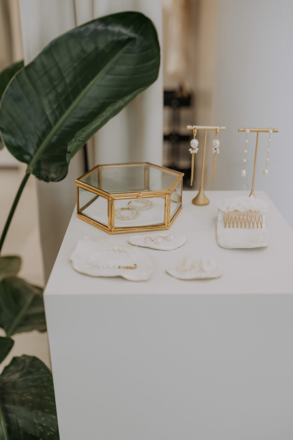 Gold- und Perlenschmuck, präsentiert auf Tabletts und Ständern auf einem weißen Sockel, neben großen grünen Blättern.