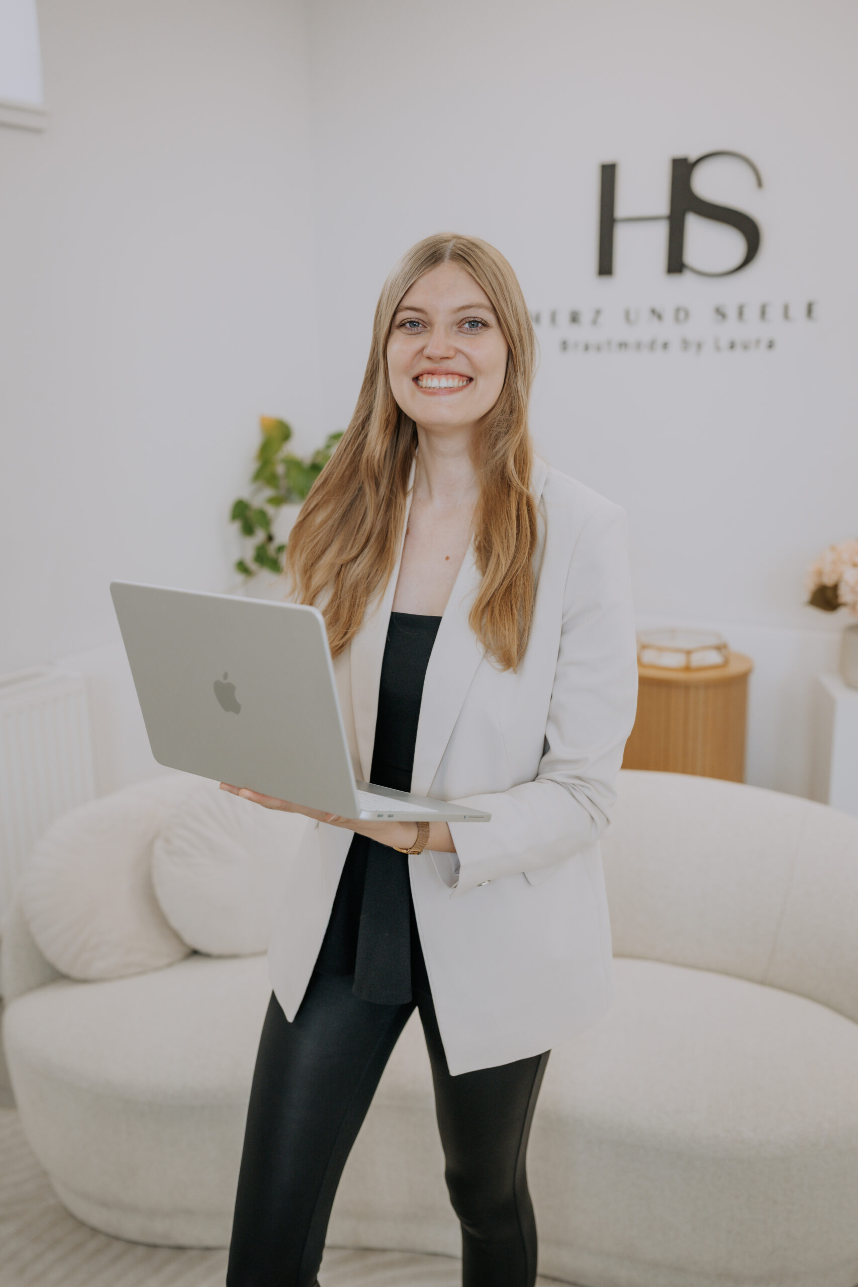 Eine lächelnde Frau in einem weißen Blazer hält einen Laptop in der Hand und steht in einem modernen, hellen Büro.