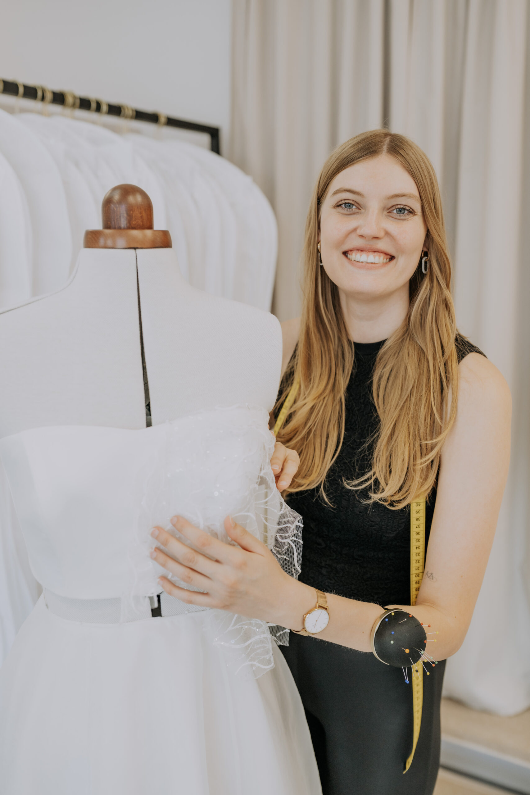 Eine lächelnde Frau, die ein weißes Kleid an einer Schaufensterpuppe zurechtzieht, mit einem Maßband um den Hals.