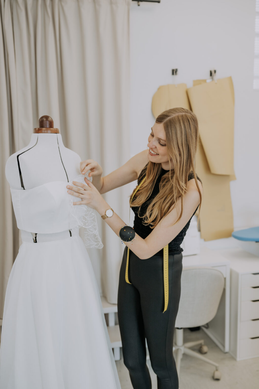 Eine Frau passt ein weißes Kleid an einer Schaufensterpuppe in einem hellen Nähatelier an, an dessen Wänden Schnittmuster hängen.