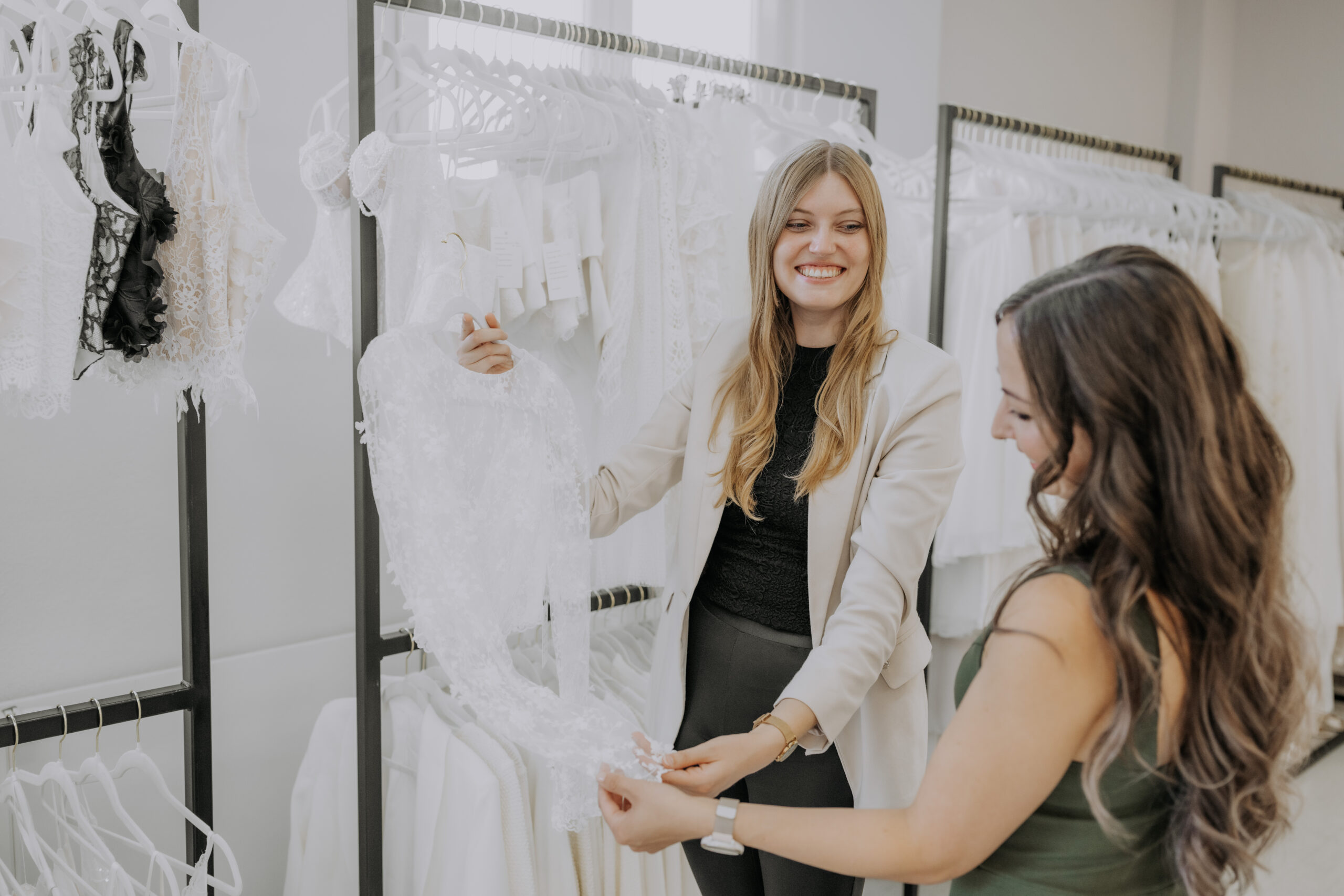 Zwei Frauen lächeln, während sie in einem Brautmodengeschäft voller Hochzeitskleider ein weißes Spitzenkleid betrachten.