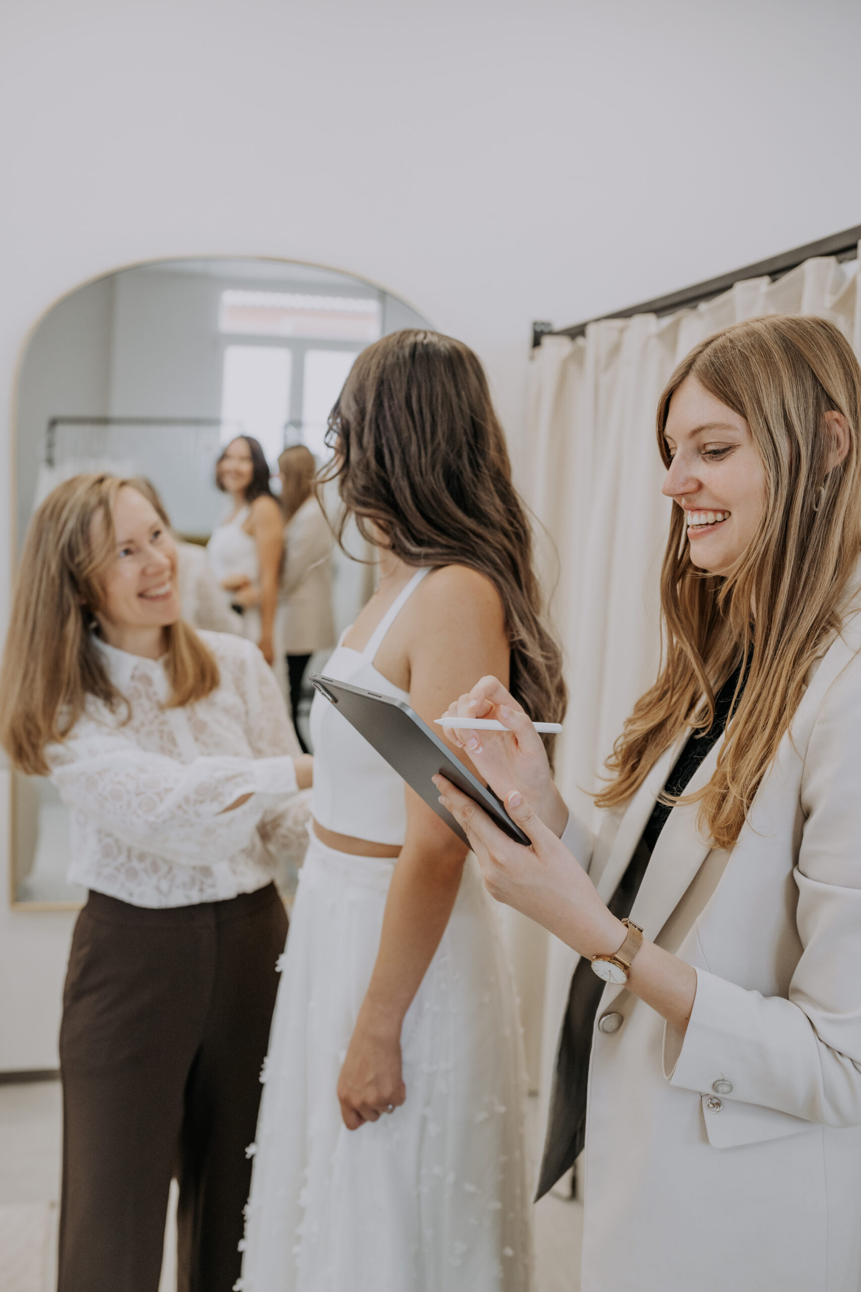 Zwei Frauen helfen einer Braut in einer Boutique; eine macht sich Notizen, während die Braut ein Brautkleid anprobiert.