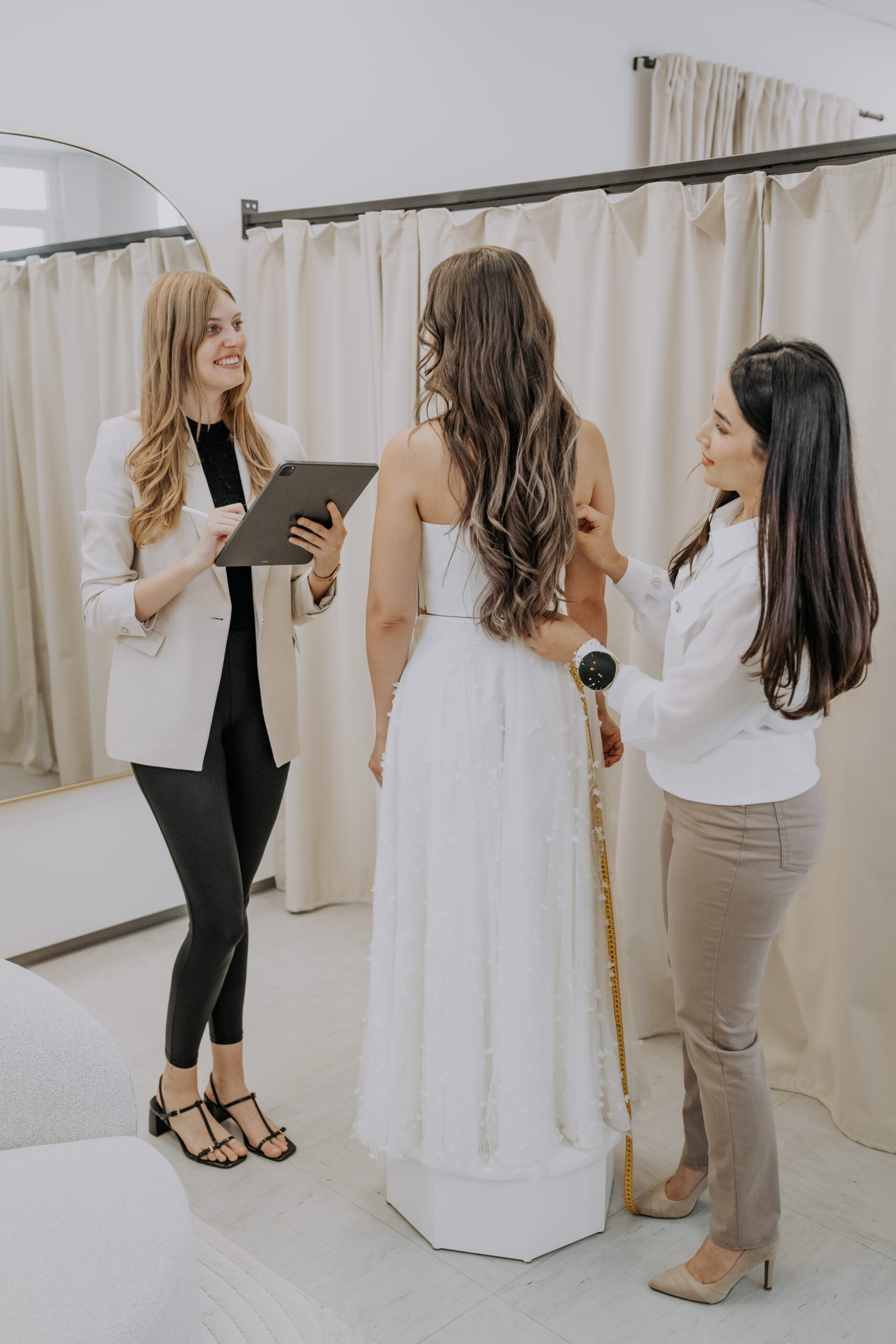 Zwei Frauen helfen einer Frau in einem weißen Kleid in einer Umkleidekabine beim Ausmessen.
